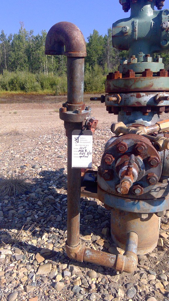 Rustic metal piping and valve equipment in an industrial setting, with a sign attached indicating 'Shut-in well' status, signifying a non-operational oil and gas well site, possibly awaiting servicing or inspection.