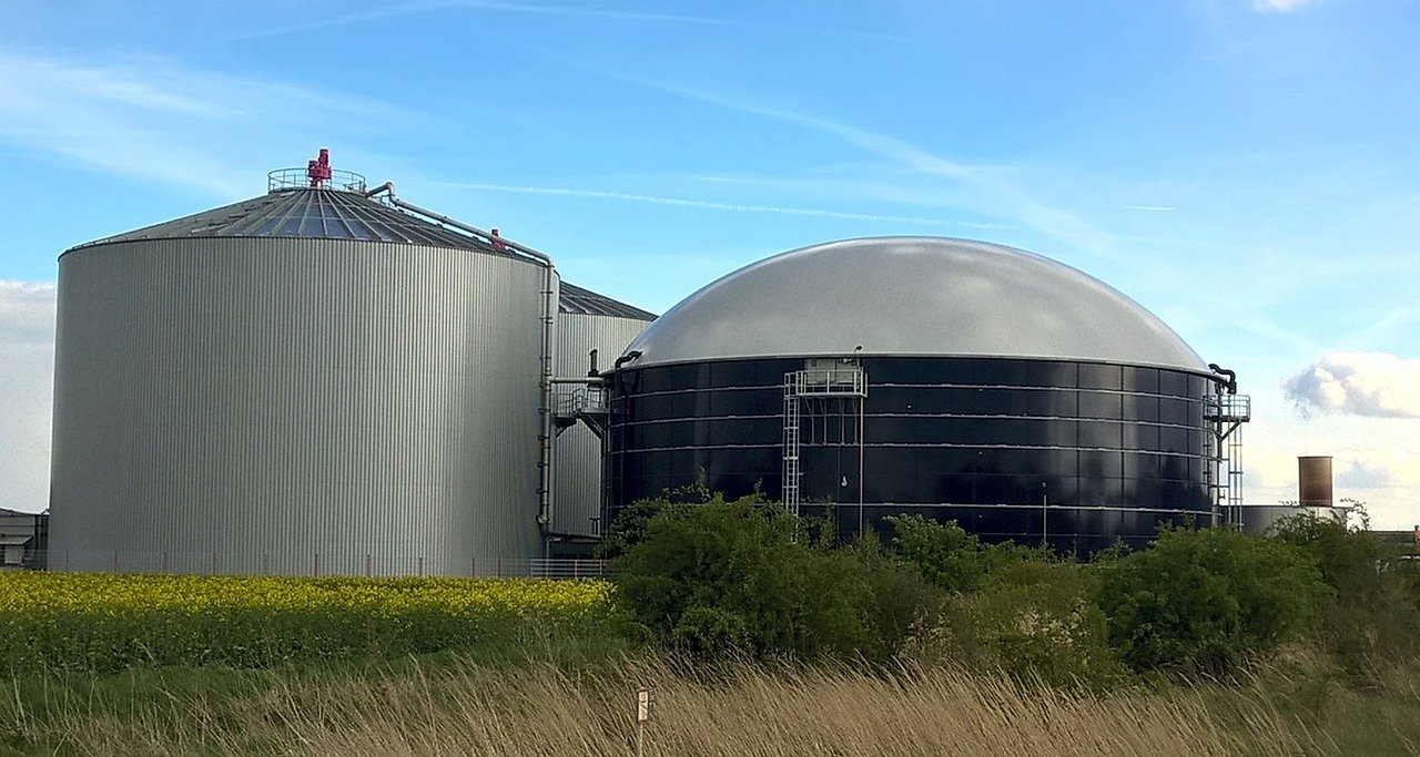Industrial biogas plants with cylindrical and dome-shaped digesters against a clear sky, highlighting renewable energy production and efforts to reduce greenhouse gas emissions.