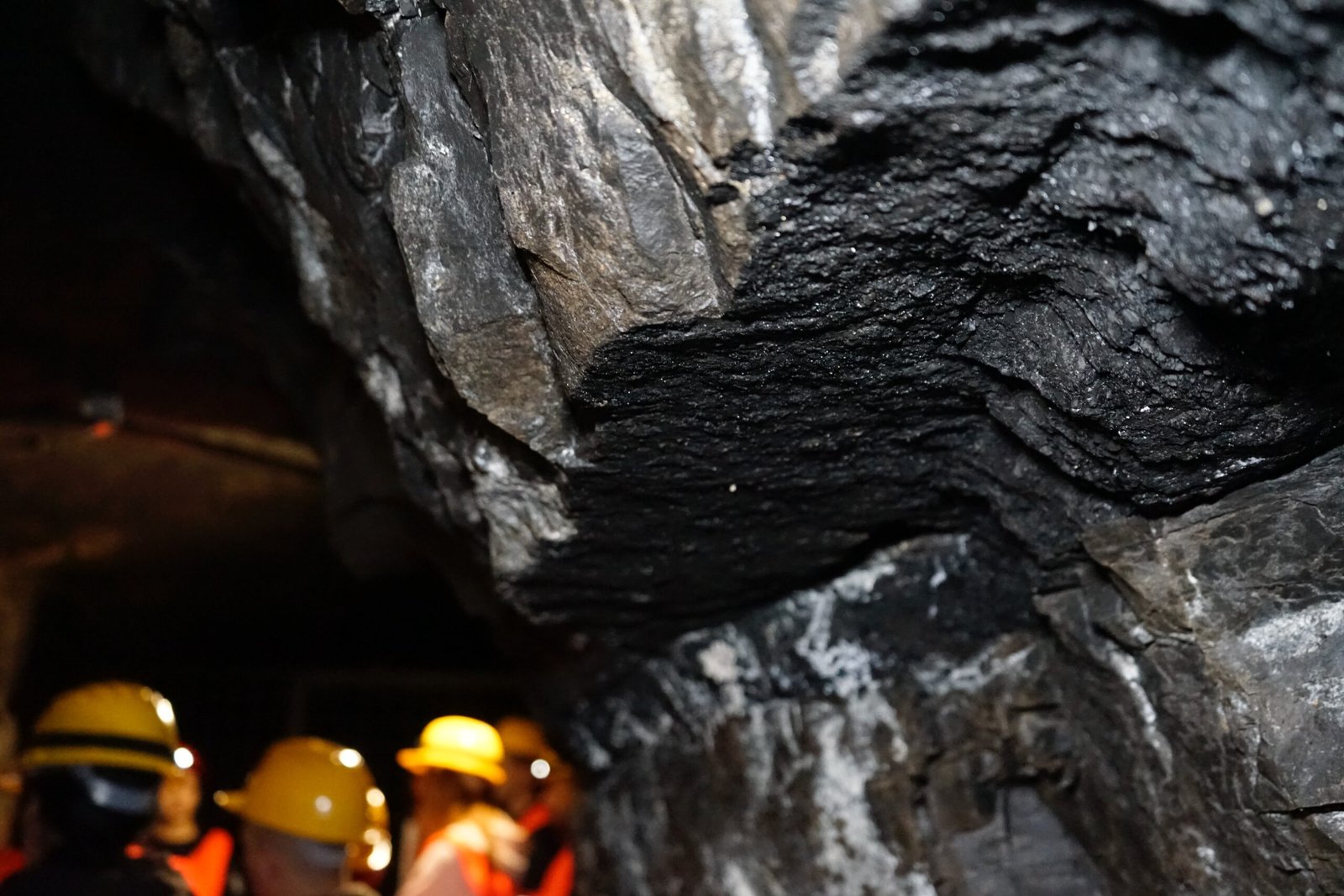 Close-up photo of coal seams in an underground mine, with the out-of-focus silhouettes of miners wearing safety helmets in the background, highlighting the extraction of fossil fuels and the workers who are integral to the coal industry.
